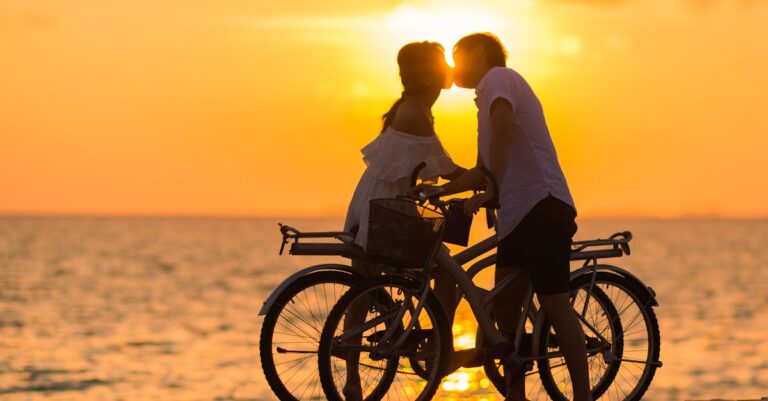 Photography of Man Wearing White T-shirt Kissing a Woman While Holding Bicycle on River Dock during Sunset