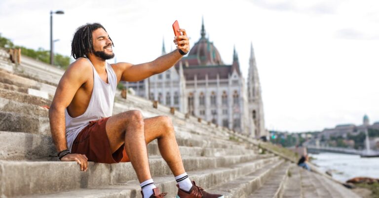 Selective Focus Photo of Smiling Man in White Vest and Red Shorts Taking a Selfie While Sitting on Concrete Stairs