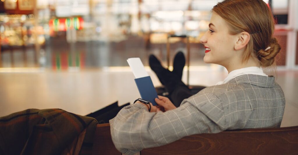 Side view of cheerful female student in checkered jacket smiling away while chilling in hall with outstretched legs and passport in hands