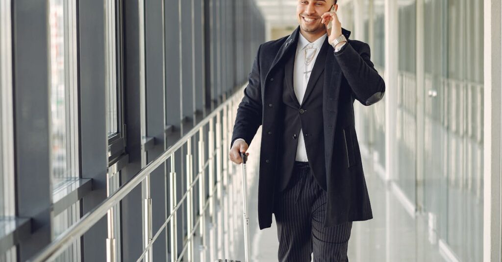 Happy ethnic male in formal suit standing in modern hallway while holding suitcase in hand and talking on mobile phone