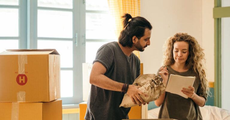 Cheerful young man and woman smiling while unpacking carton boxes with belongings in new apartment during relocation and looking at paper