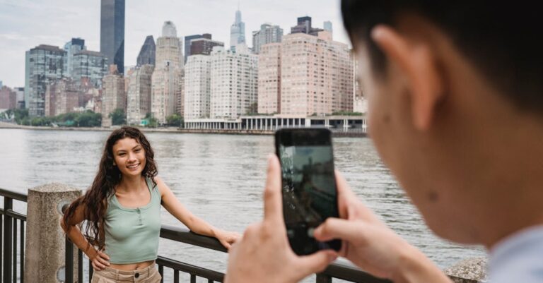 Crop unrecognizable young guy using smartphone while photographing happy young ethnic girlfriend standing on river embankment with hand on waist during trip in New York City