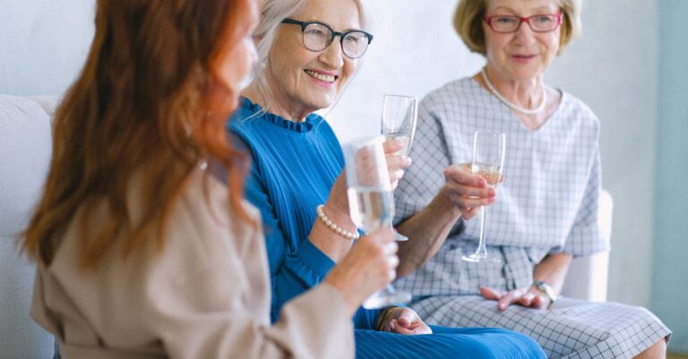 Elderly cheerful women with glasses of champagne smiling and chatting while celebrating special festive event