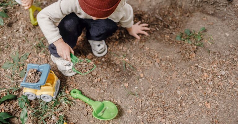 High angle of little kid in brown hat squatting on ground and playing with plastic colourful toys