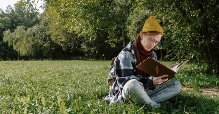 Woman in Blue Denim Jacket and Yellow Knit Cap Sitting on Green Grass Field Reading Book