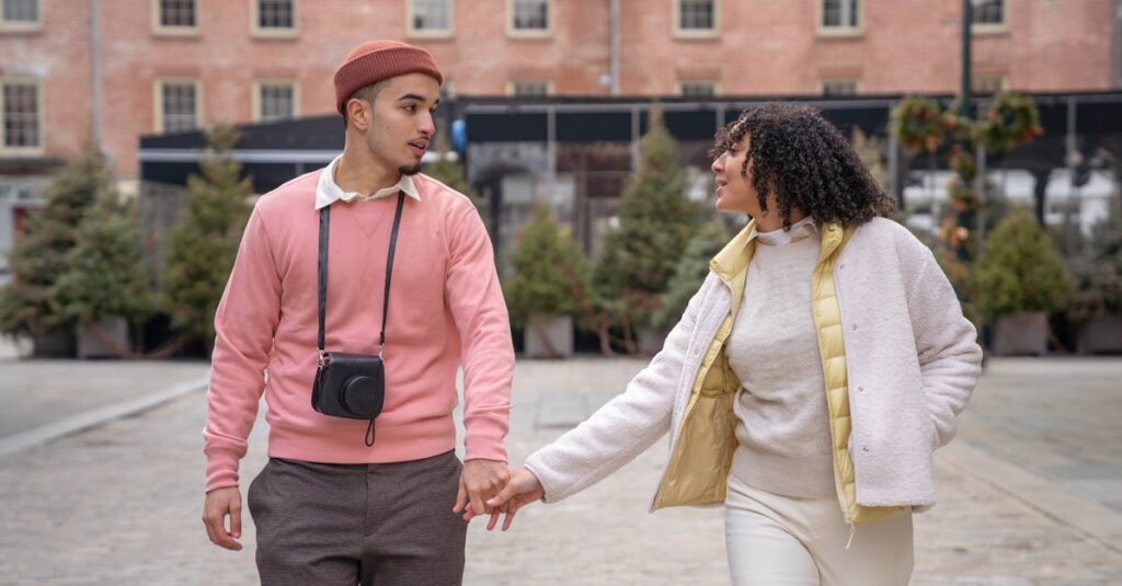 Positive young ethnic couple in stylish warm clothes holding hands while walking in aged district of city during vacation and looking at each other