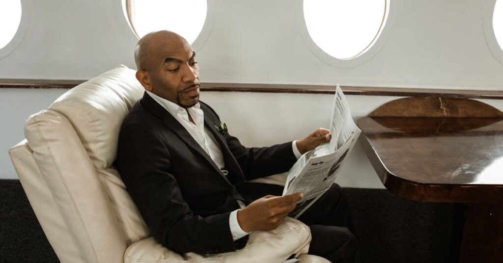 An adult man in a black suit reads a newspaper while sitting in a private jet's luxurious interior.