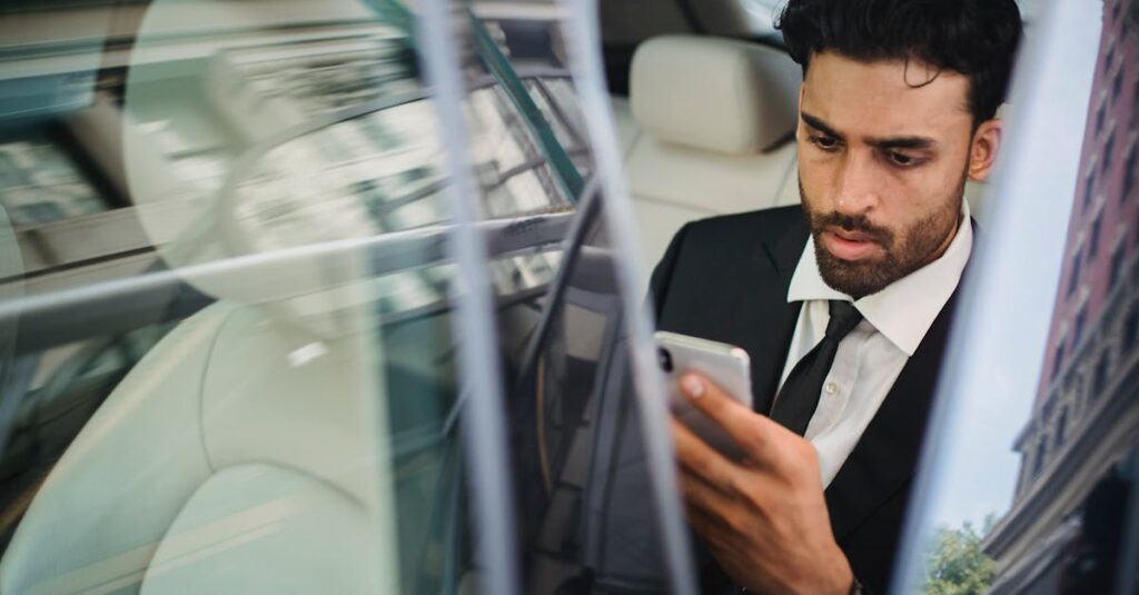 Confident businessman in suit using smartphone inside car, viewed through window reflection.