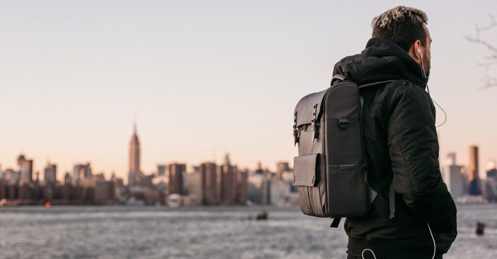 A man wearing a backpack gazes at the Manhattan skyline from Brooklyn waterfront during the day.