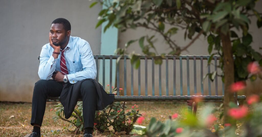 A businessman in a suit sits pensively on a bench in a tranquil park setting.