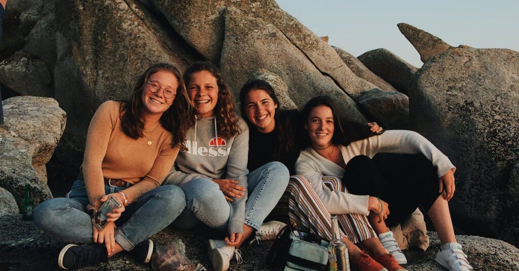 Group of cheerful young female friends in casual clothes laughing and looking at camera while sitting with drinks and snacks on rocky seashore and having fun during travel together