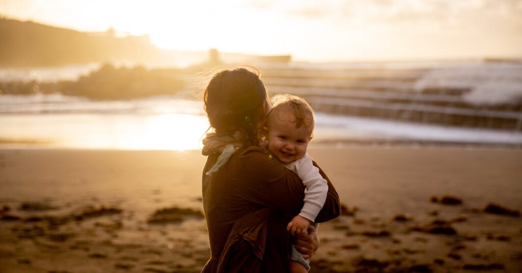 A tender moment of a mother holding her smiling baby at the beach during sunset.